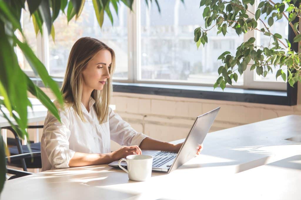 Beautiful Caucasian Girl Working On A Laptop Remotely In A Bright Space With Green Plants.