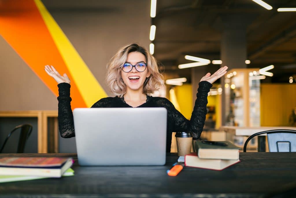 Funny Happy Excited Young Pretty Woman Sitting At Table In Black Shirt Working On Laptop In Co Working Office, Wearing Glasses